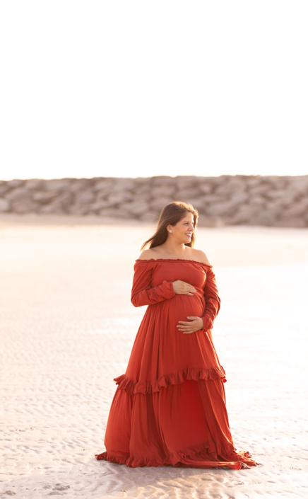 regnant woman standing near the waterline at Al Khan Beach in Sharjah during a maternity photoshoot, softly holding her baby bump.