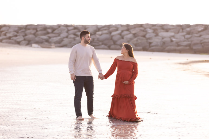 Pregnant woman sitting on the sand near rocks at Al Mamzar Beach during a maternity photoshoot, looking out over the sea