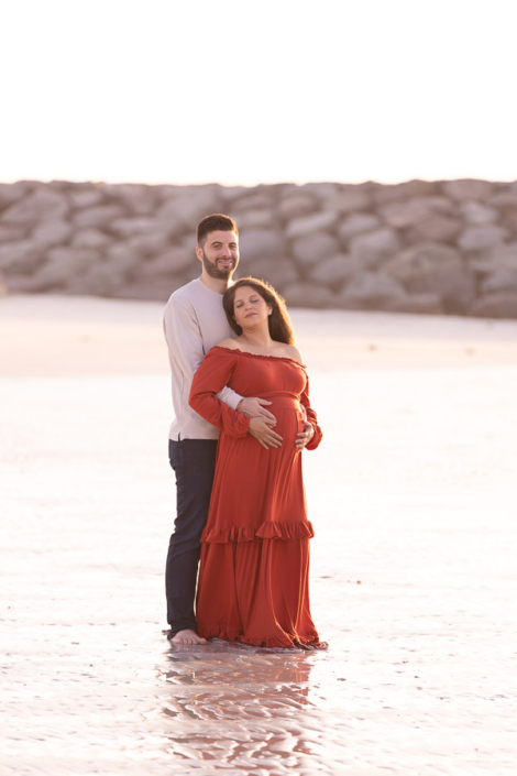 Pregnant woman standing on a quiet beach at sunset in the UAE during a maternity photoshoot, with warm light and gentle waves around her
