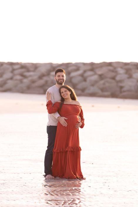 Expecting couple standing close together at Al Mamzar Beach between Dubai and Sharjah during a maternity photoshoot, with the sea and city towers in the distance