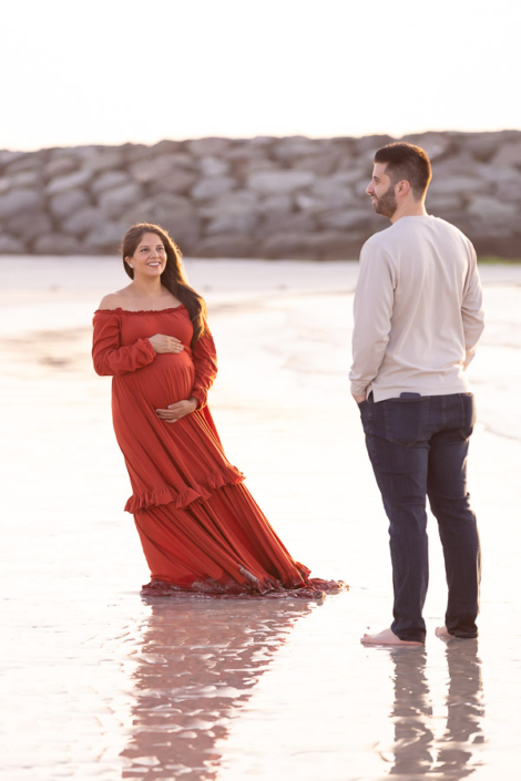 Pregnant mother walking towards the camera along Jumeirah Beach in Dubai, smiling as the waves roll in behind her.
