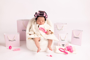 A newborn baby girl wearing a tiny white spa bathrobe and a head towel, resting in a luxury white setup with miniature designer-style bags in a Dubai villa.
